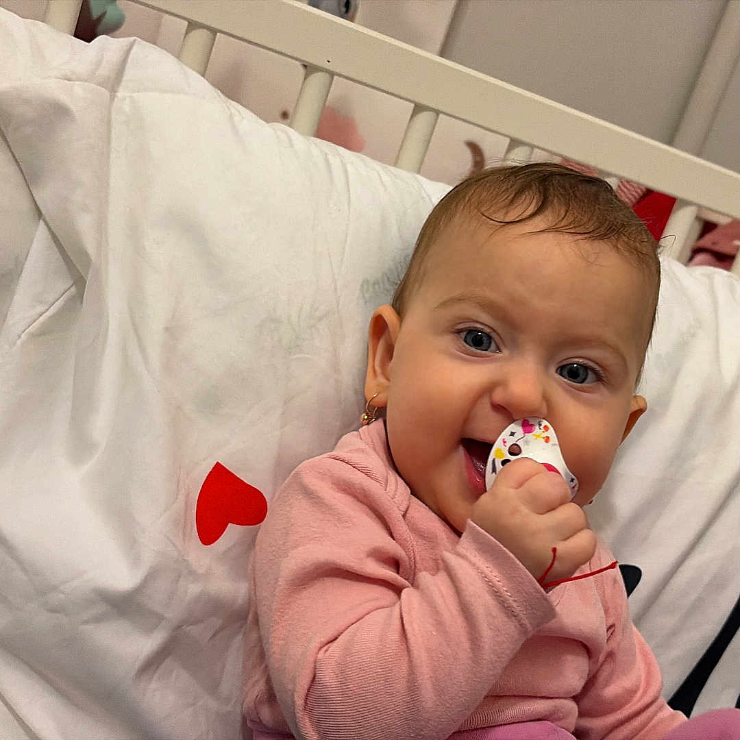 Aryana-Maria participe au concours pour gagner de l'argent avec cette photo : baby, baby_toy, child, cozy, crib, cute, earrings, face, happy, head, indoor, infant, pacifier, person, pink_clothing, portrait, red_hearts, sitting, smile, white_bedding