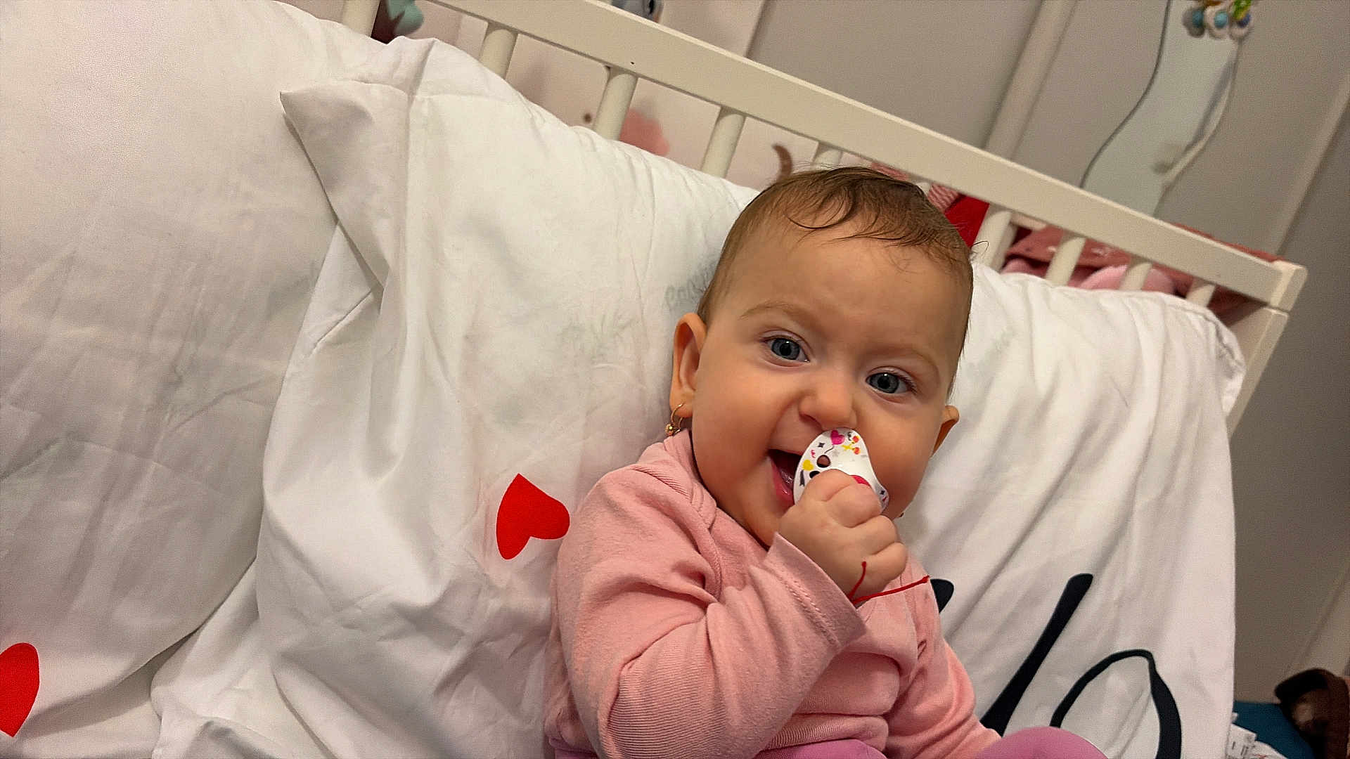 Aryana-Maria participe au concours pour gagner de l'argent avec cette photo : baby, infant, pacifier, pink_clothing, crib, white_bedding, red_hearts, earrings, smile, happy, child, indoor, cute, portrait, person, baby_toy, head, face, sitting, cozy