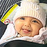 baby, infant, child, smiling, tongue_out, portrait, close_up, winter_clothing, knit_hat, pom_pom, pink_clothing, scarf, car_seat, seat_belt, vehicle_interior, warning_label, outdoor_travel, face, happy, photography