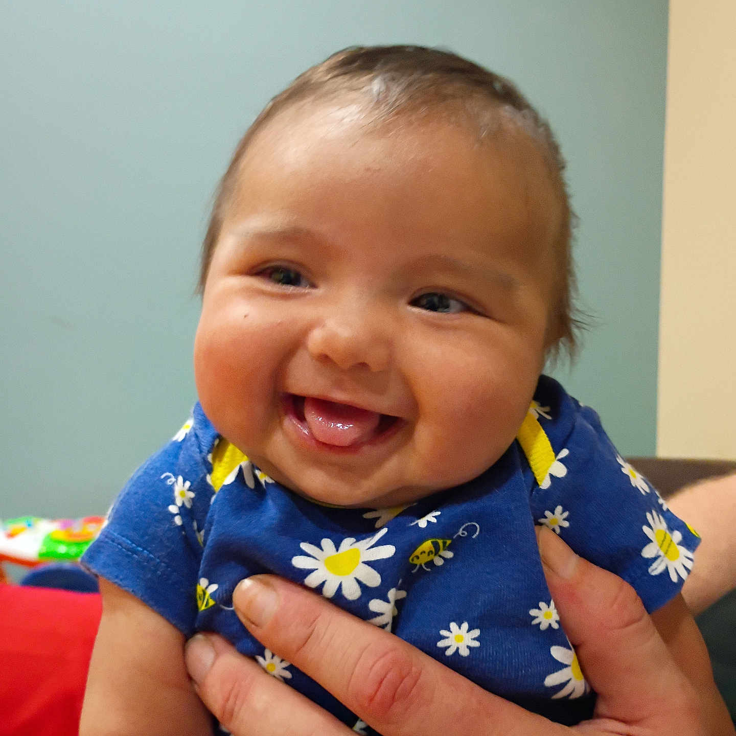 Rylann joined the competition — help win amazing prizes! adult_hand, baby, background, blue_clothing, cheeks, child, closeup, cute, face, floral_pattern, hand, happy, holding, indoor, infant, person, portrait, skin, smiling, tongue