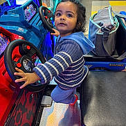 Rishabh is registered to the contest to win money with this photo: toddler, child, arcade, driving_game, steering_wheel, blue_sweater, indoor, play, game, colorful_lights, bag, seat, floor, red, blue, curious, young_child, toy, fun, entertainment