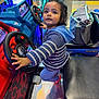 toddler, child, arcade, driving_game, steering_wheel, blue_sweater, indoor, play, game, colorful_lights, bag, seat, floor, red, blue, curious, young_child, toy, fun, entertainment