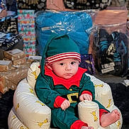 Billy-James is registered to the contest to win money with this photo: baby, child, elf_costume, holiday, christmas, gift, present, wrapped_gift, plush_chair, giraffe_pattern, indoors, footwear, package, curious, cute, festive, holiday_season, childrens_clothing, sitting, expression