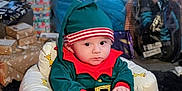 Billy-James is registered to the contest to win money with this photo: baby, child, childrens_clothing, christmas, curious, cute, elf_costume, expression, festive, footwear, gift, giraffe_pattern, holiday, holiday_season, indoors, package, plush_chair, present, sitting, wrapped_gift