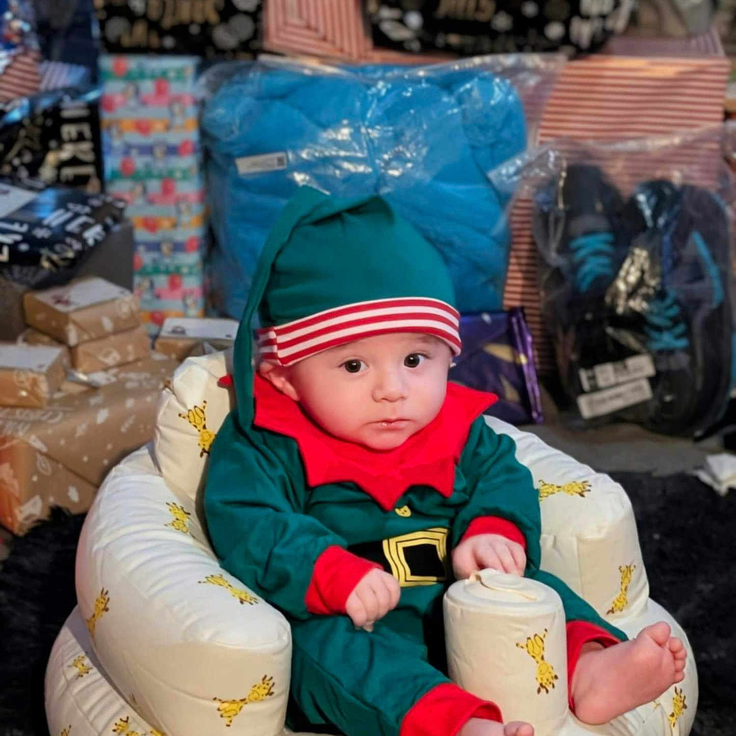 Billy-James is registered to the contest to win money with this photo: baby, child, childrens_clothing, christmas, curious, cute, elf_costume, expression, festive, footwear, gift, giraffe_pattern, holiday, holiday_season, indoors, package, plush_chair, present, sitting, wrapped_gift