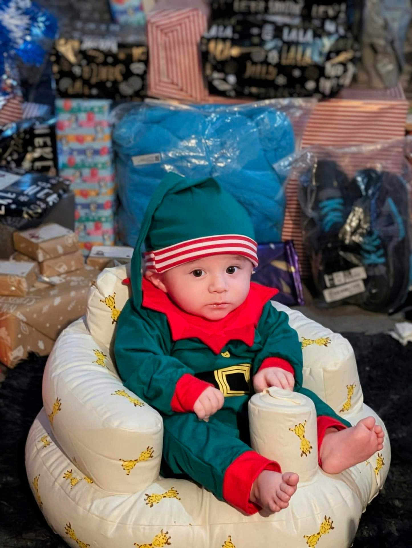 Billy-James is registered to the contest to win money with this photo: baby, child, elf_costume, holiday, christmas, gift, present, wrapped_gift, plush_chair, giraffe_pattern, indoors, footwear, package, curious, cute, festive, holiday_season, childrens_clothing, sitting, expression