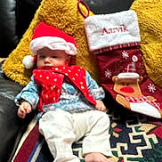 Aarvik is registered to the contest to win money with this photo: baby, infant, santa_hat, red_vest, white_pants, couch, yellow_pillow, christmas_stocking, holiday, festive, indoors, carpet, barefoot, child, person, furniture, relaxing, decor, seasonal, cozy
