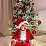 baby, child, santa_claus_costume, christmas_tree, holiday, festive, lights, ornaments, red_clothing, hat, blanket, indoors, decorations, celebration, cute, smiling, seasonal, warm, cozy, portrait