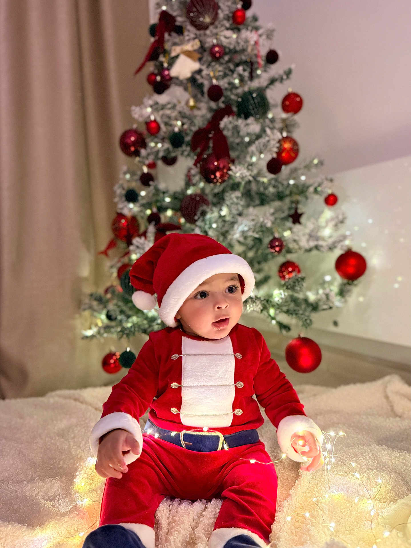 Joud participe au concours pour gagner de l'argent avec cette photo : baby, child, santa_claus_costume, christmas_tree, holiday, festive, lights, ornaments, red_clothing, hat, blanket, indoors, decorations, celebration, cute, smiling, seasonal, warm, cozy, portrait