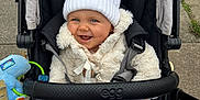 Beau joined the competition — help win amazing prizes! baby, stroller, white_hat, pom_pom, blanket, smile, child, outdoor, jacket, happy, cute, baby_toy, person, face, cold_weather, fur_coat, seat, black, blue_eyes, pavement