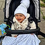 Beau is registered to the contest to win money with this photo: baby, child, stroller, white_hat, pom_pom, blanket, toy, smiling, outdoor, pavement, clothing, bundle_up, cute, happy, face, person, cold_weather, baby_jacket, baby_accessory, infant