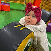Beau is registered to the contest to win money with this photo: baby, child, indoor_play_area, padded_block, colorful, pink_bow, fluffy_coat, curious_expression, soft_mats, netting, playground, toddler, headband, cartoon_eye, green_floor, orange_walls, purple_block, yellow_poles, childrens_play_area, person