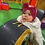 baby, child, indoor_play_area, padded_block, colorful, pink_bow, fluffy_coat, curious_expression, soft_mats, netting, playground, toddler, headband, cartoon_eye, green_floor, orange_walls, purple_block, yellow_poles, childrens_play_area, person