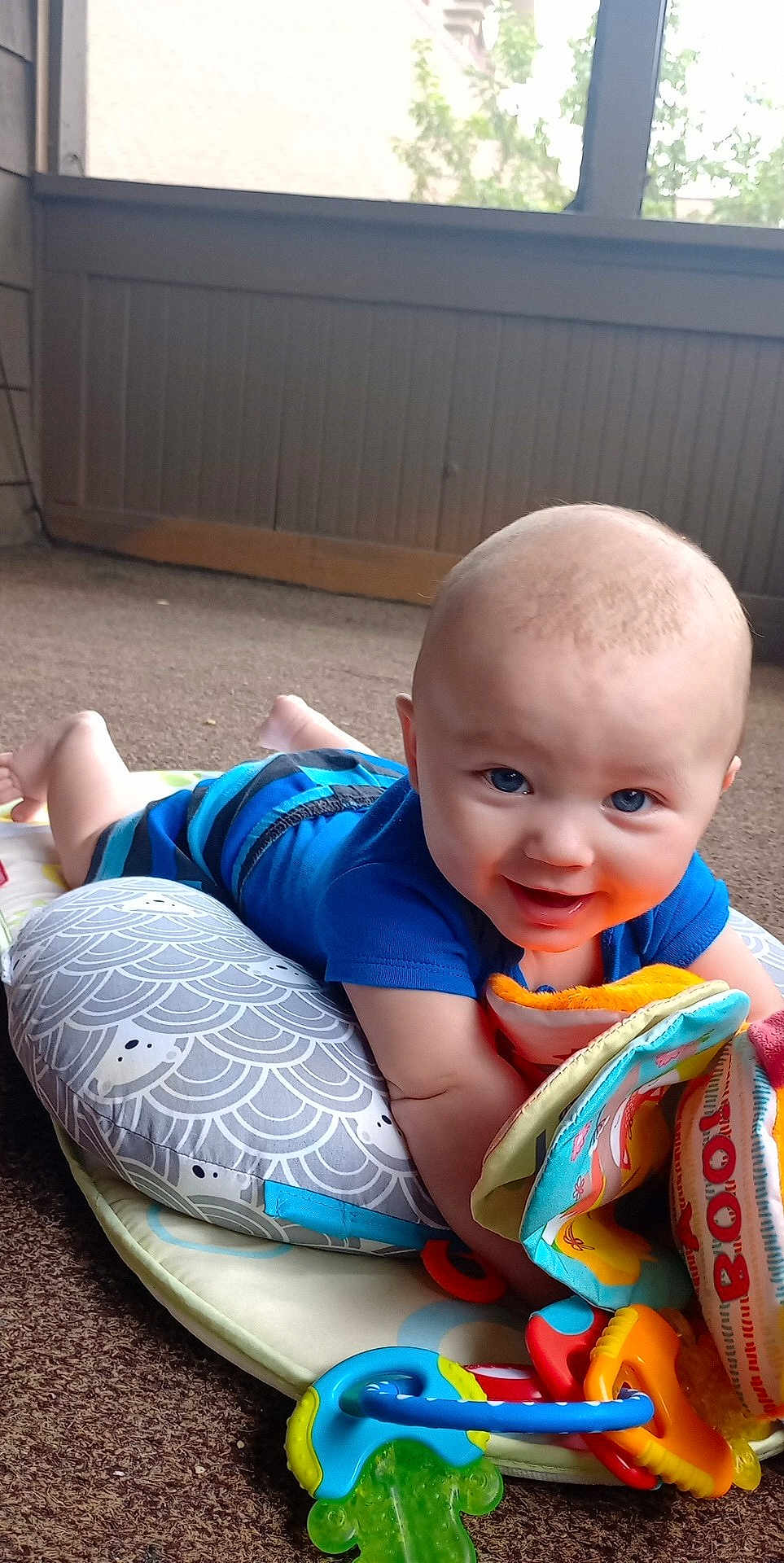 Abel is registered to the contest to win money with this photo: baby, infant, smiling, toy, play_mat, soft_book, pillow, indoor, carpet, child, blue_clothing, colorful, cute, happy, face, person, floor, playtime, young_child, toddler