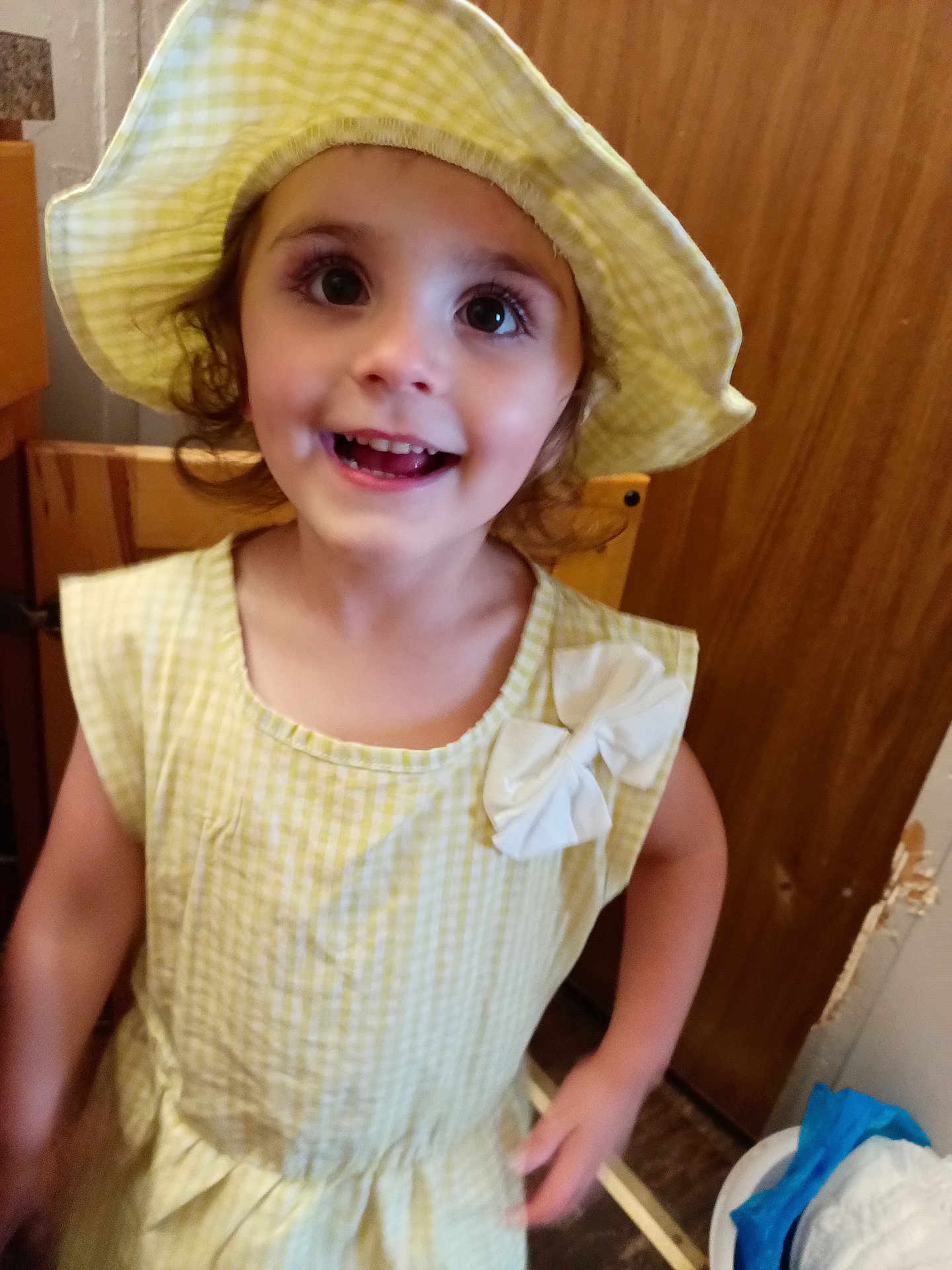 Alora is registered to the contest to win money with this photo: child, girl, dress, hat, yellow, smiling, happy, indoor, portrait, cute, young, face, person, clothing, curly_hair, warm_lighting, wood, home, standing, bow