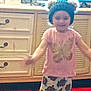 child, indoor, hat, pajamas, cabinet, furniture, smile, girl, colorful, patterned_clothing, blurred, overexposed, happy, standing, home, decor, red_cushions, wood_floor, face, person