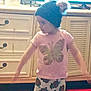 Alora is registered to the contest to win money with this photo: child, indoors, cabinet, drawer, television, clothing, beanie, pants, tshirt, butterfly, patterned_pants, pom_poms, standing, floor, red_cushions, furniture, blurry, grainy, person, home