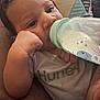Masai is registered to the contest to win money with this photo: baby, bottle, drinking, infant, close_up, face, hand, clothing, patterned_pillow, curly_hair, milk, resting, contemplative, cute, indoor, person, laying, young_child, soft_light, casual