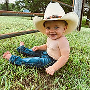 Crew is registered to the contest to win money with this photo: toddler, child, cowboy_hat, barefoot, grass, jeans, smiling, outdoor, fence, nature, greenery, happy, portrait, baby, sitting, sunlight, rural, cute, person, casual