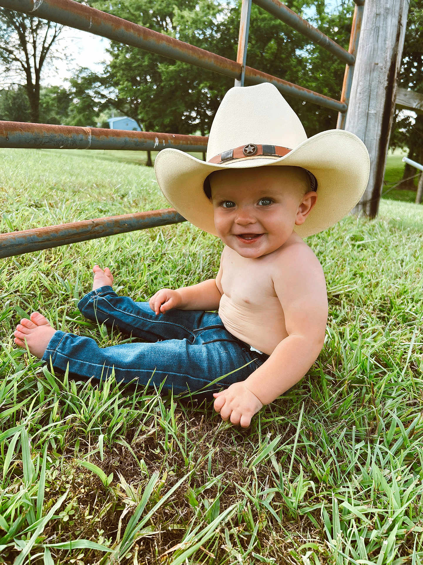 Crew is registered to the contest to win money with this photo: toddler, child, cowboy_hat, barefoot, grass, jeans, smiling, outdoor, fence, nature, greenery, happy, portrait, baby, sitting, sunlight, rural, cute, person, casual
