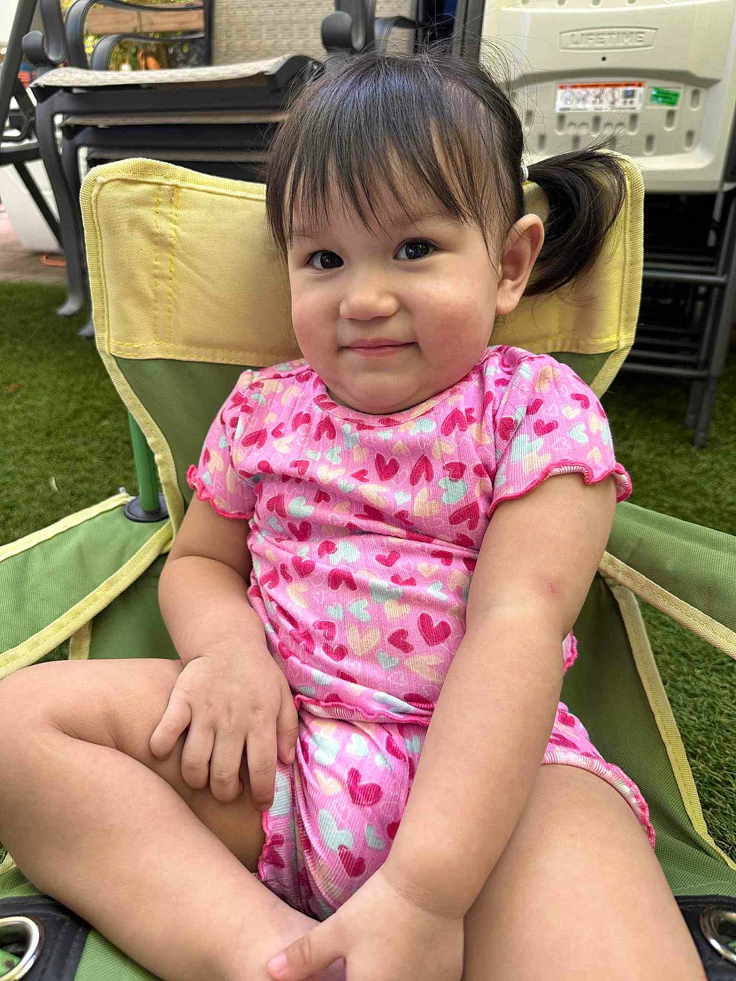 Angelina is registered to the contest to win money with this photo: toddler, child, smiling, pink_clothing, heart_pattern, chair, outdoor, grass, short_hair, pigtails, cute, sitting, happy, young_child, casual, daylight, skin, person, relaxed, summer