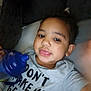 Malakai is registered to the contest to win money with this photo: toddler, child, face, expression, sippy_cup, blue, shirt, text, lying_down, cushion, indoors, cozy, cute, person, casual_clothing, young_child, playful, smile, hand, relaxed
