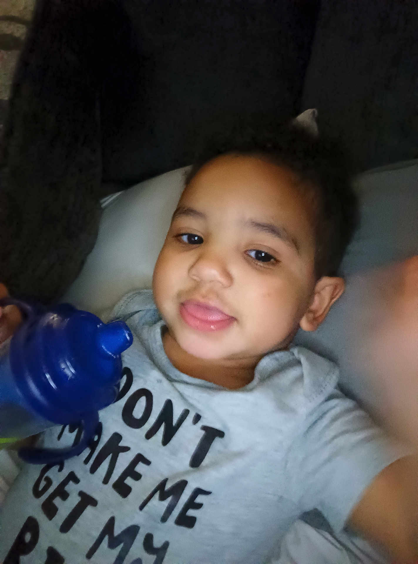 Malakai is registered to the contest to win money with this photo: toddler, child, face, expression, sippy_cup, blue, shirt, text, lying_down, cushion, indoors, cozy, cute, person, casual_clothing, young_child, playful, smile, hand, relaxed