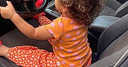 Inaya participe au concours pour gagner de l'argent avec cette photo : toddler, child, car_interior, steering_wheel, curly_hair, orange_shirt, patterned_pants, car_seat, dashboard, window, door, side_mirror, seatbelt, car_door, vehicle, driving, playful, indoor, young_child, automobile