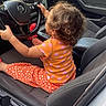 toddler, child, car_interior, steering_wheel, curly_hair, orange_shirt, patterned_pants, car_seat, dashboard, window, door, side_mirror, seatbelt, car_door, vehicle, driving, playful, indoor, young_child, automobile