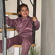 Inaya participe au concours pour gagner de l'argent avec cette photo : child, smiling, purple_outfit, stairs, spiral_stairs, wooden_stairs, metal_railings, indoor, cozy, happy, person, casual_clothing, wall, plant, home_interior, footwear, socks, young, portrait, relaxed