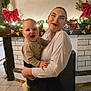 baby, woman, child, smile, happy, holding, indoor, fireplace, holiday_decor, christmas_lights, red_bow, face, person, clothing, floor, tile, home, celebration, joy, family