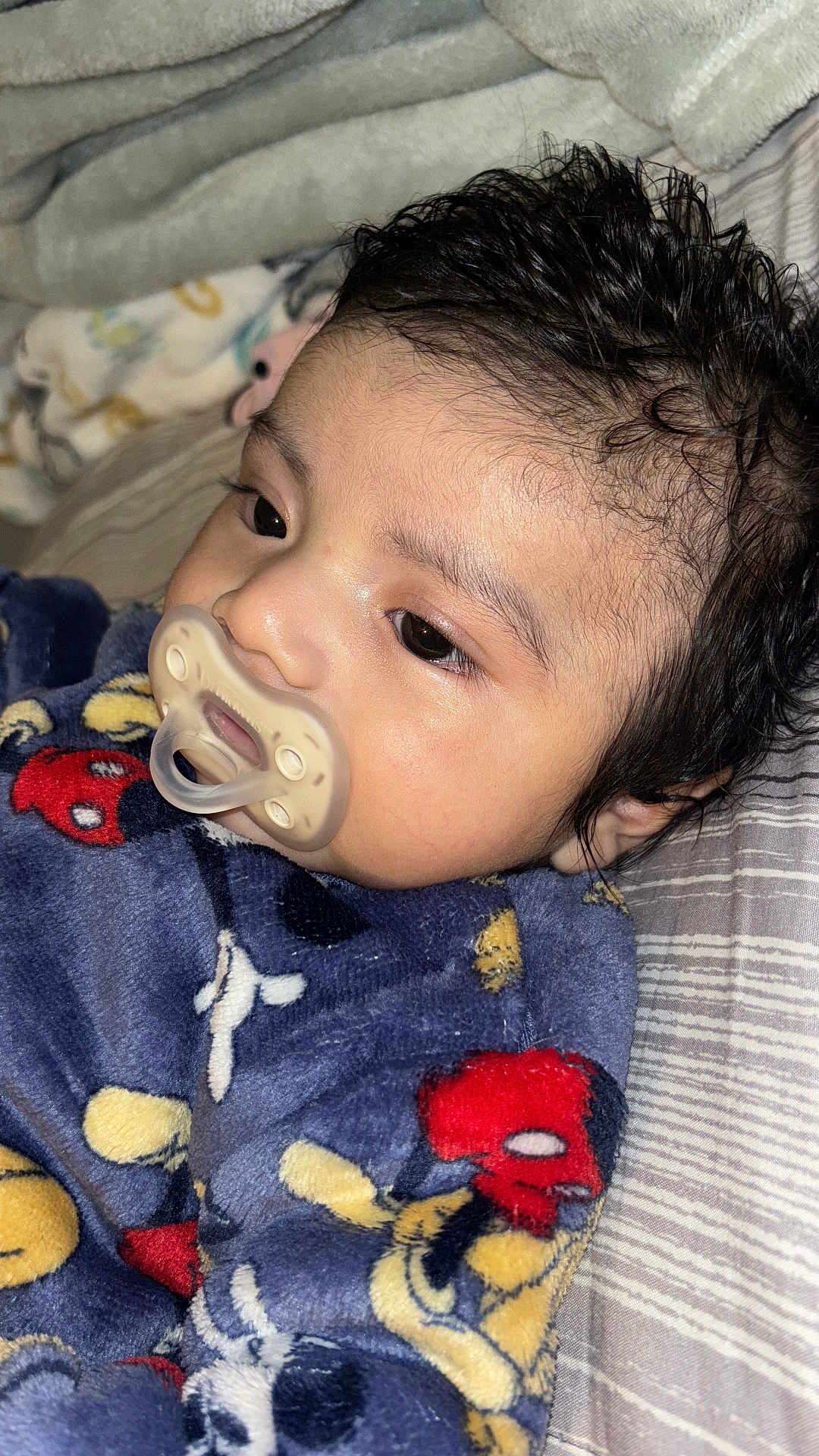 Ian is registered to the contest to win money with this photo: baby, pacifier, blanket, mickey_mouse, bed, striped_bedspread, curly_hair, infant, close_up, sleepwear, soft_texture, indoors, resting, child, young_child, face, head, cozy, comfort, cute