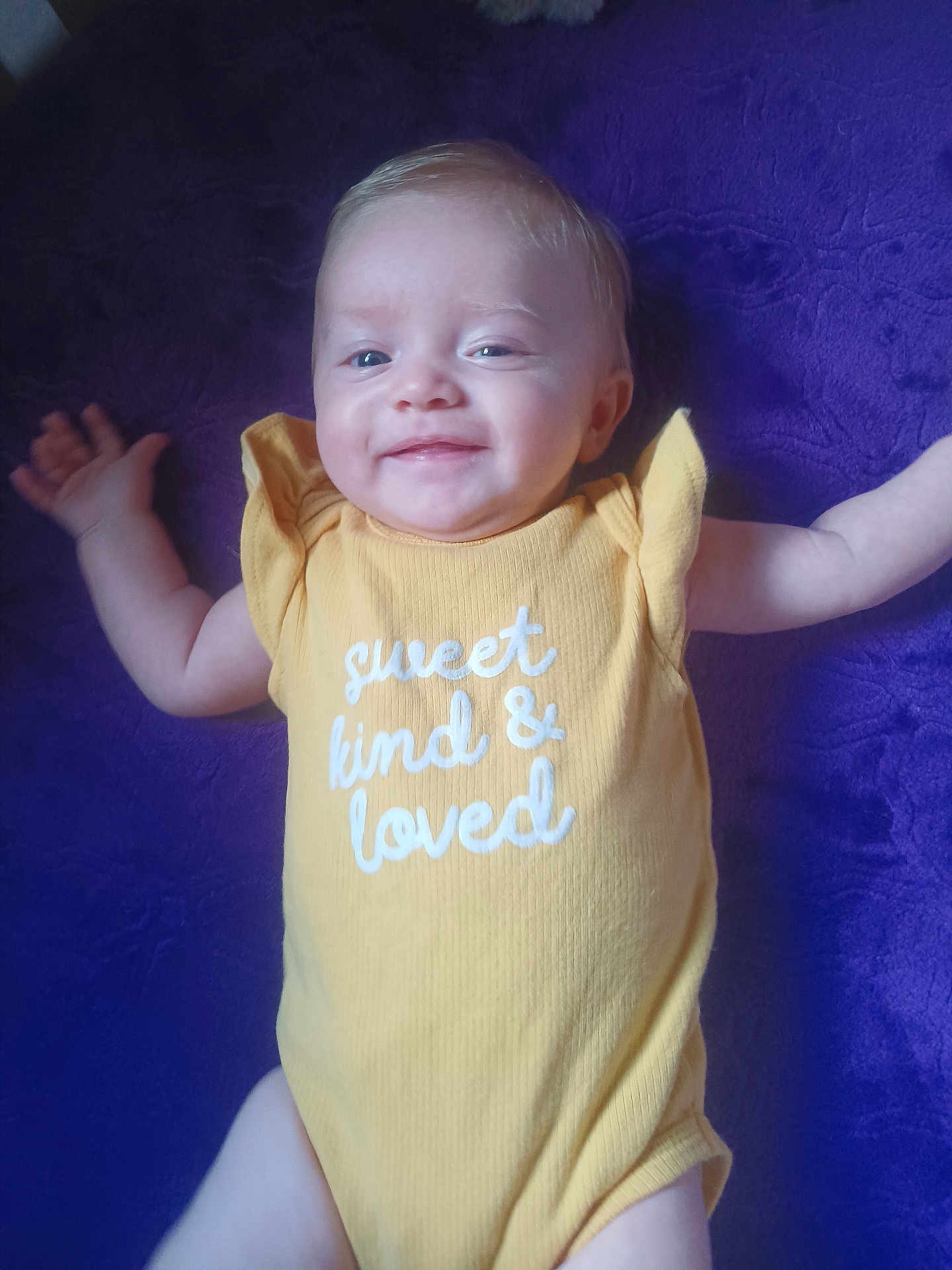 Lainey is registered to the contest to win money with this photo: baby, smiling, yellow_onesie, purple_blanket, infant, happy, face, arms, cute, child, lying_down, text_on_clothing, portrait, skin, one_person, indoors, young_child, soft_texture, fabric, human