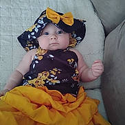 Zelpa is registered to the contest to win money with this photo: baby, child, hat, bow, floral_clothing, yellow_skirt, quilt, couch, indoor, portrait, cute, infant, person, fashion, sitting, smile, skin, face, arm, legs