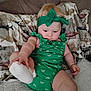 baby, child, green_outfit, headband, sock, couch, blanket, indoor, curious, sitting, infant, cute, plush, patterned_blanket, soft_lighting, arm, leg, foot, face, person