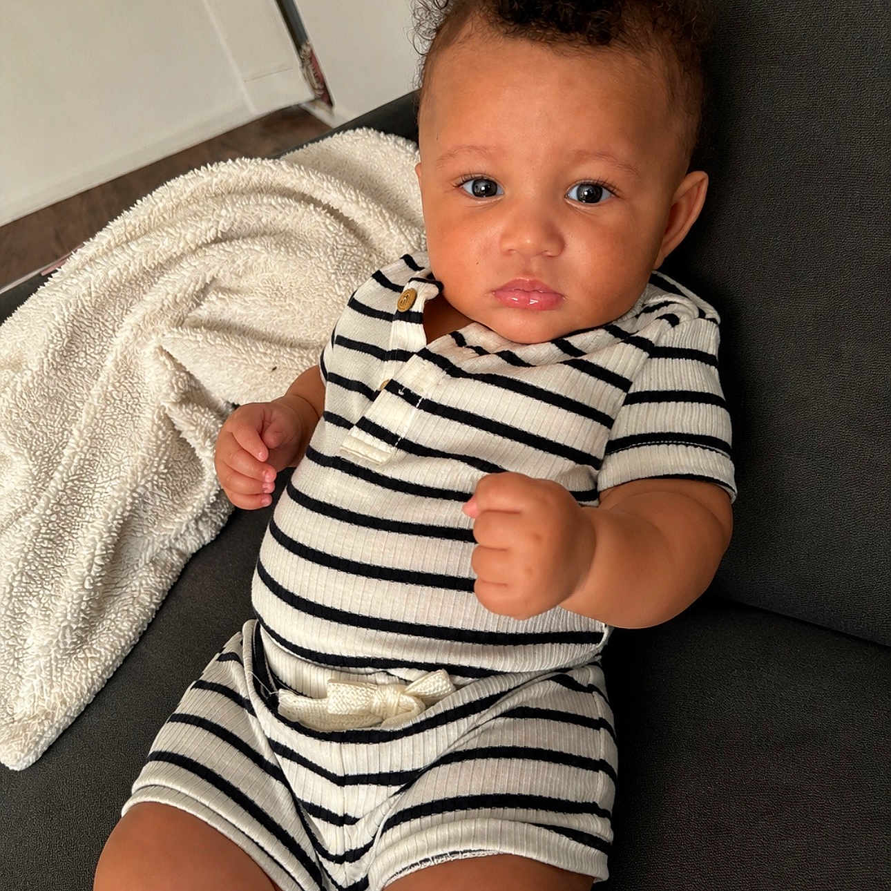 Carter is registered to the contest to win money with this photo: baby, child, curly_hair, striped_clothing, couch, blanket, indoors, portrait, person, furniture, cute, infant, expression, raised_fists, seated, cozy, soft_texture, casual, young, looking_at_camera