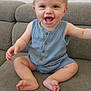 baby, child, smile, happy, sitting, romper, blue_clothing, couch, furniture, indoor, cute, infant, face, skin, arm, foot, toes, eyes, head, person