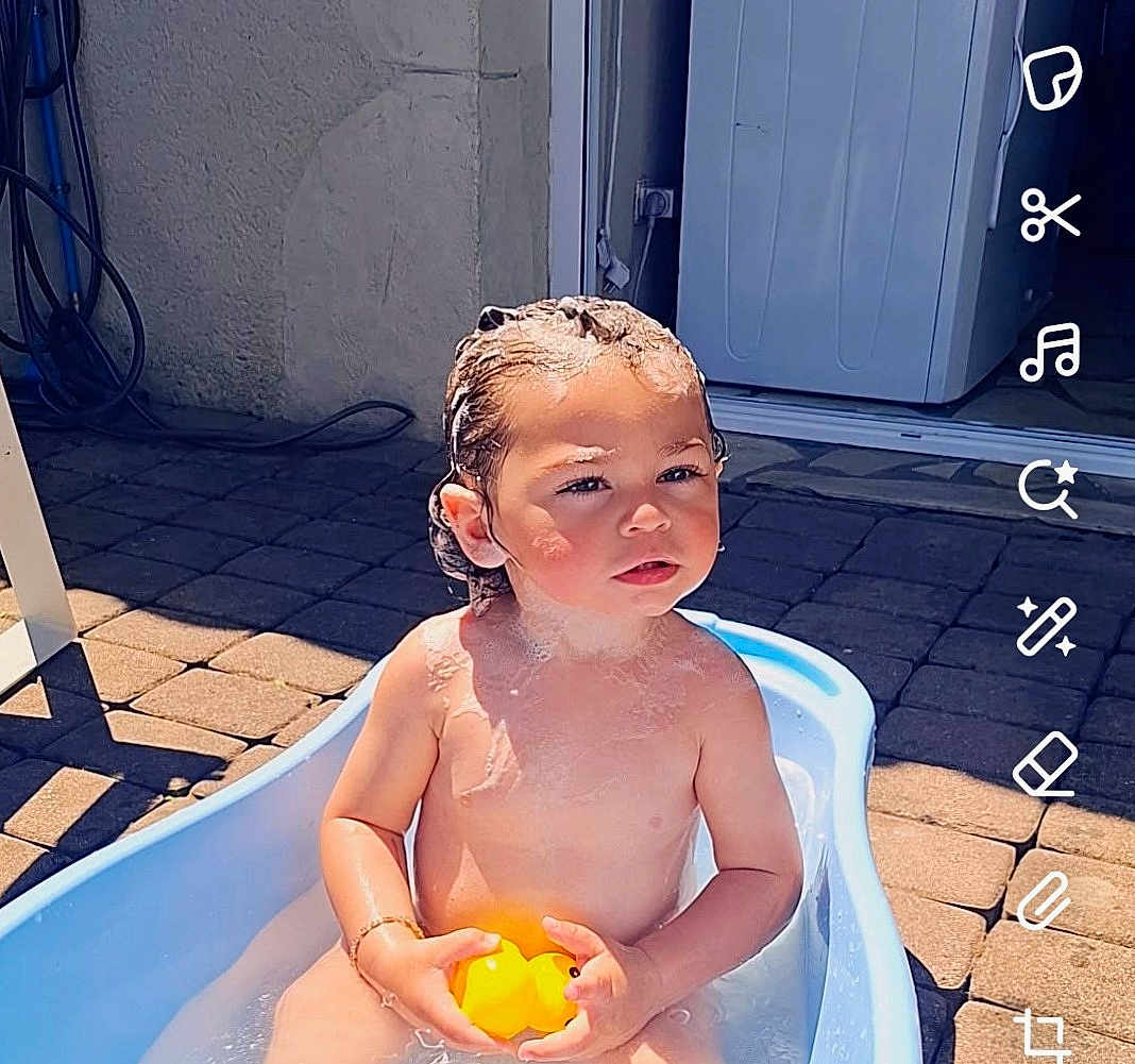 Nayden a rejoint le concours — aidez-le/la à gagner de superbes lots ! baby, bath_time, bathtub, child, curious_expression, daylight, door, outdoor, pavement, person, playful, rubber_duck, skin, soap_suds, summer, sunlight, toddler, toy, water, wet_hair
