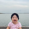 baby, smiling, child, headband, denim, water, lake, outdoor, happy, sitting, infant, toddler, casual_clothing, nature, sky, cloudy, portrait, cute, joyful, accessory