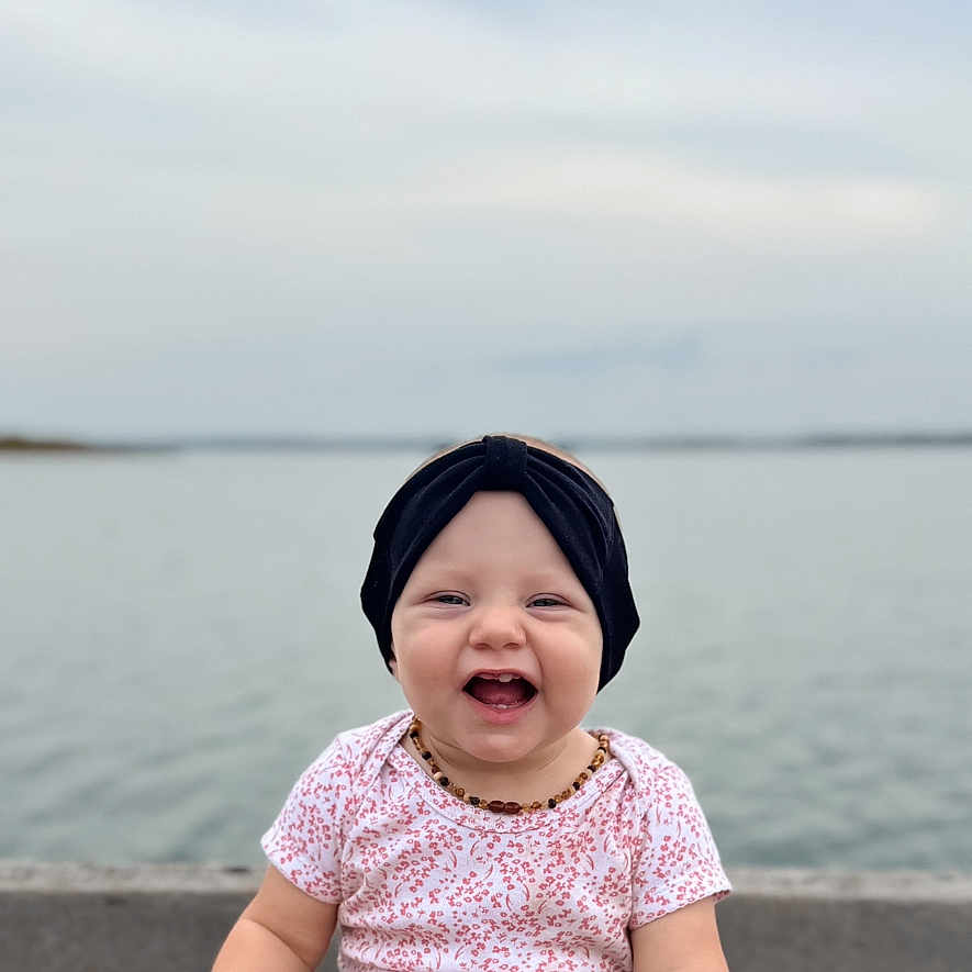 Makenzie joined the competition — help win amazing prizes! baby, smiling, child, headband, denim, water, lake, outdoor, happy, sitting, infant, toddler, casual_clothing, nature, sky, cloudy, portrait, cute, joyful, accessory