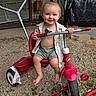toddler, child, smile, tricycle, outdoor, barefoot, gravel, red, toy, play, summer, happy, person, shorts, shirt, small_hands, topknot, vehicle, seat, handlebars