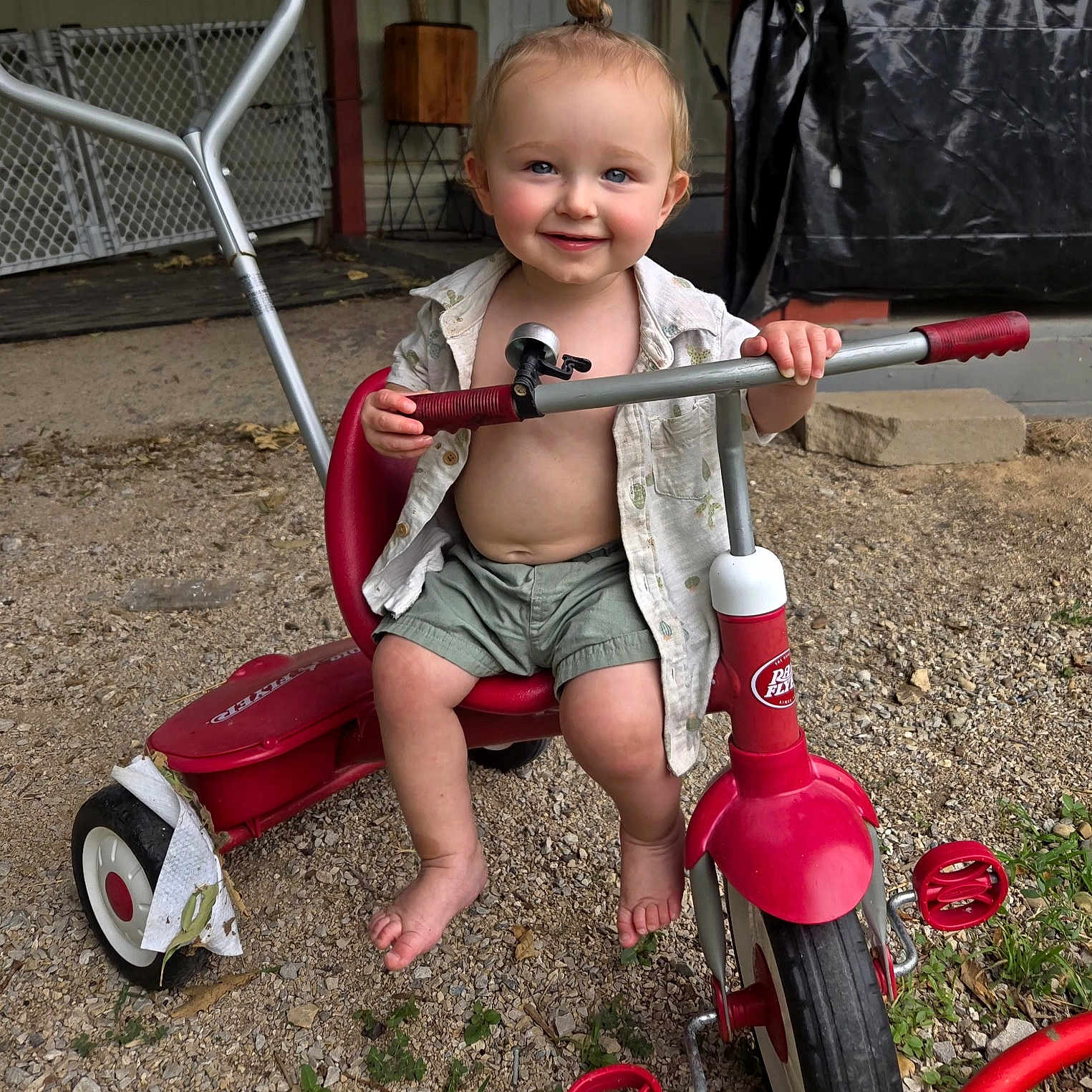 Brewski is registered to the contest to win money with this photo: toddler, child, smile, tricycle, outdoor, barefoot, gravel, red, toy, play, summer, happy, person, shorts, shirt, small_hands, topknot, vehicle, seat, handlebars
