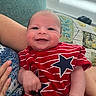 baby, smiling, infant, red_shirt, stars, striped_clothing, arm, hand, couch, quilt, fabric, person, indoor, portrait, happy, closeup, skin, head, face, child