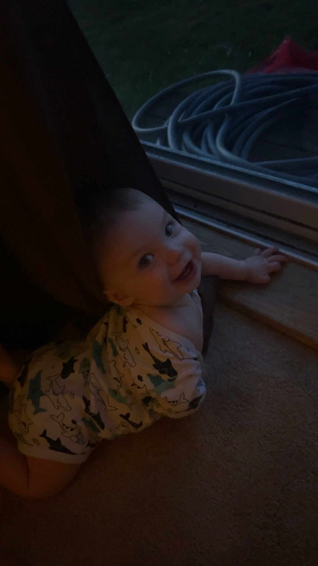 Emmett is registered to the contest to win money with this photo: baby, infant, crawling, onesie, shark_pattern, curtain, door, glass_door, hose, indoor, carpet, smiling, child, face, hand, window, dark, cozy, floor, home
