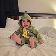 Skylar Scott is registered to the contest to win money with this photo: baby, child, dinosaur_costume, onesie, green_clothing, hood, smiling, laughing, sitting, bed, bedroom, pillow, blanket, bare_feet, toes, portrait, indoor, cozy, happy, photography