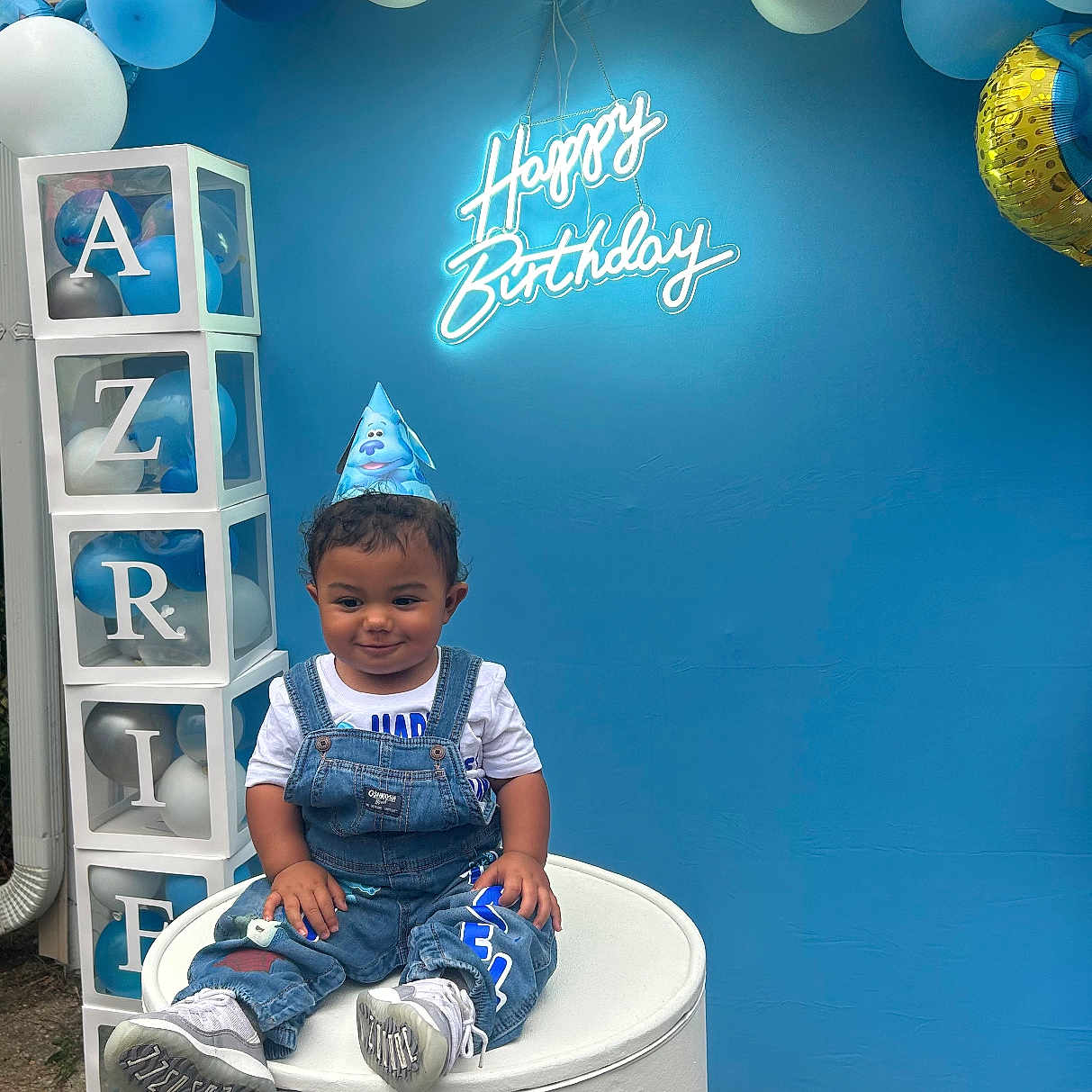 Azriel joined the competition — help win amazing prizes! balloons, birthday, blue_backdrop, blue_theme, casual_clothing, celebration, child, curly_hair, cylindrical_pedestal, decorative_blocks, denim_overalls, event, festive, happy_birthday, neon_sign, outdoor, party_hat, smiling_child, toddler, white_shoes