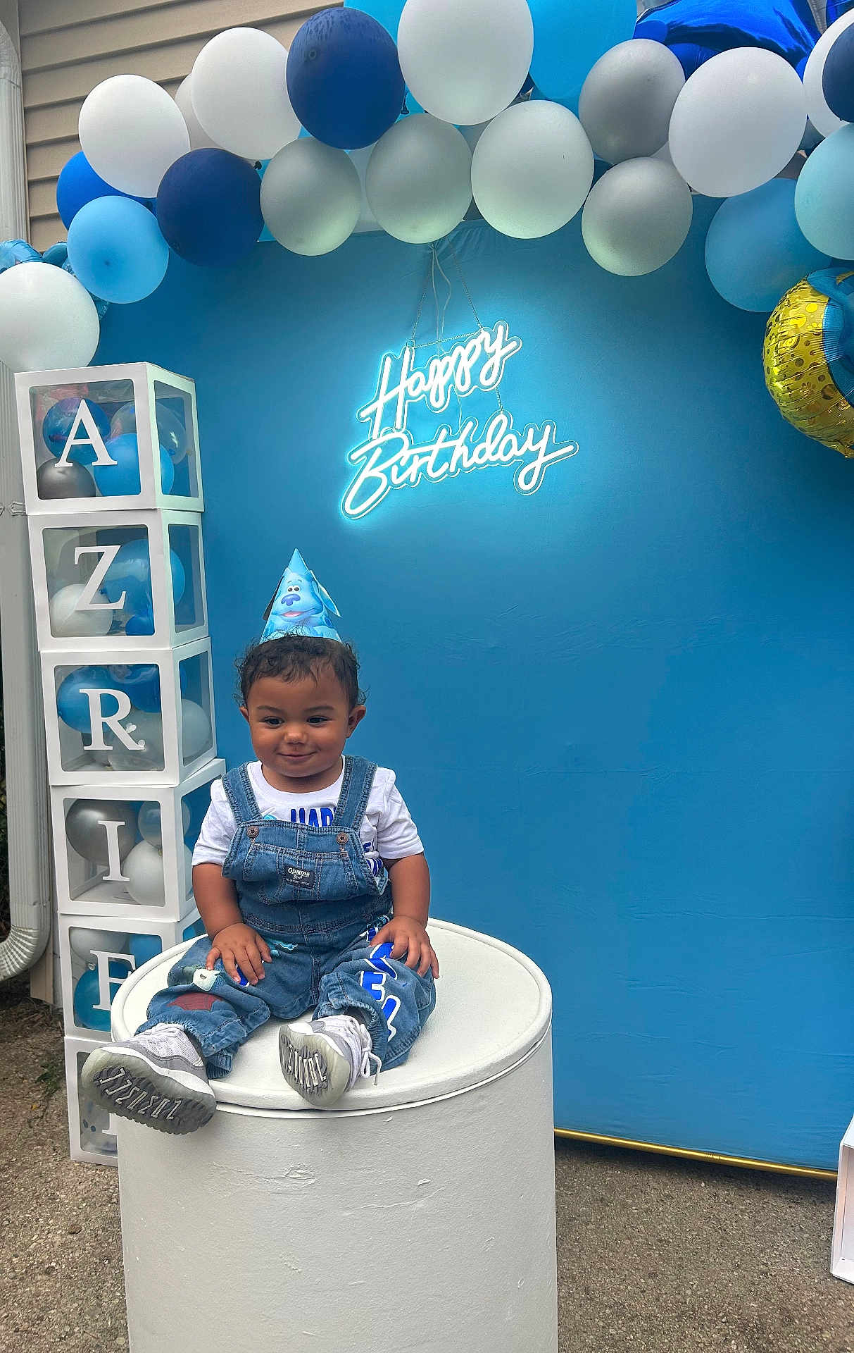 Azriel joined the competition — help win amazing prizes! balloons, birthday, blue_backdrop, blue_theme, casual_clothing, celebration, child, curly_hair, cylindrical_pedestal, decorative_blocks, denim_overalls, event, festive, happy_birthday, neon_sign, outdoor, party_hat, smiling_child, toddler, white_shoes