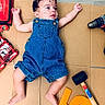baby, cardboard, casual_clothing, child, curly_hair, cute, denim_overalls, drill, expression, floor, indoor, infant, lying_down, person, playful, portrait, rubber_mallet, toolkit, tools, wood_floor