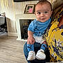 baby, child, infant, blue_clothing, socks, white_shoes, adult, floral_top, living_room, fireplace, wooden_floor, portrait, smiling, person, home_interior, casual, seated, indoor, cute, holding