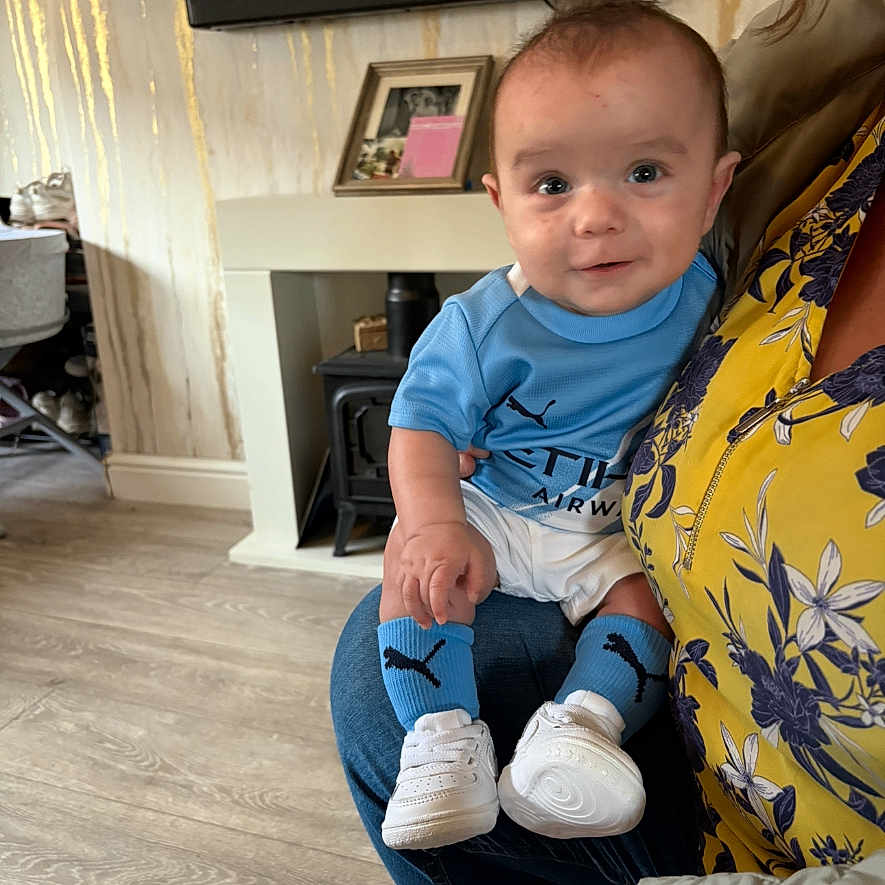 Riko joined the competition — help win amazing prizes! adult, baby, blue_clothing, casual, child, cute, fireplace, floral_top, holding, home_interior, indoor, infant, living_room, person, portrait, seated, smiling, socks, white_shoes, wooden_floor